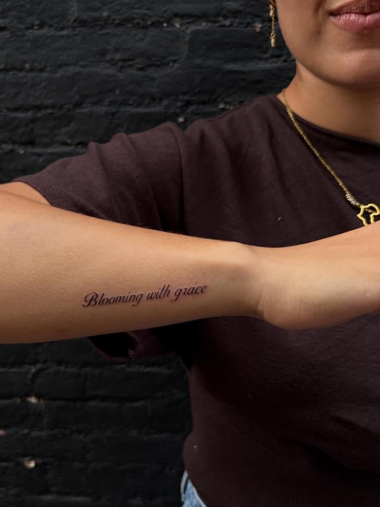 Fine-line script tattoo reading Blooming with grace on an inner forearm.