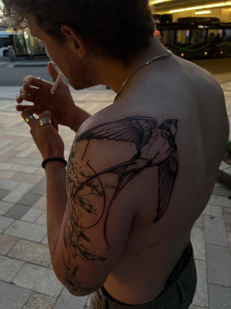 Black-and-grey swallow tattoo across a left shoulder and upper arm, photographed outdoors at night.